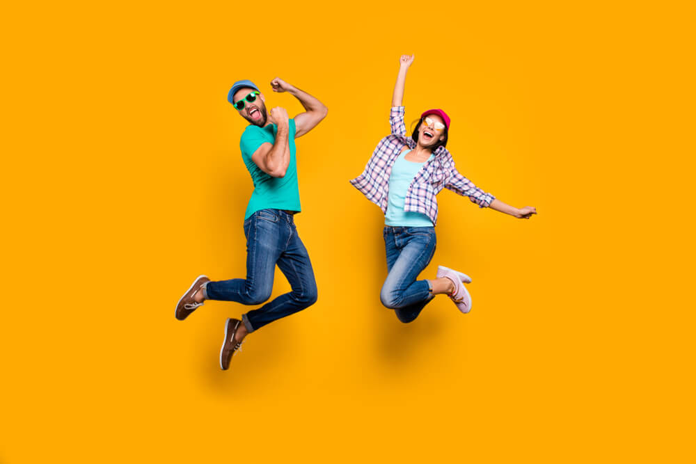 Portrait of funky active couple jumping with raised fists celebrating victory wearing casual clothes isolated on vivid yellow background; enthusiastic consent concept